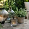 Gartendeko mit Glas auf Holztisch mit Laternen und Pflanzen im gemütlichen Gartenbereich.