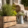 Terrassendeko mit alten Sachen aus Holzkiste, Kräutern und Gießkanne auf gemütlicher Terrasse.