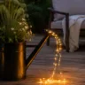 Neue Gießkanne mit LED-Lichterkette als Gartendeko auf beleuchteter Terrasse am Abend.