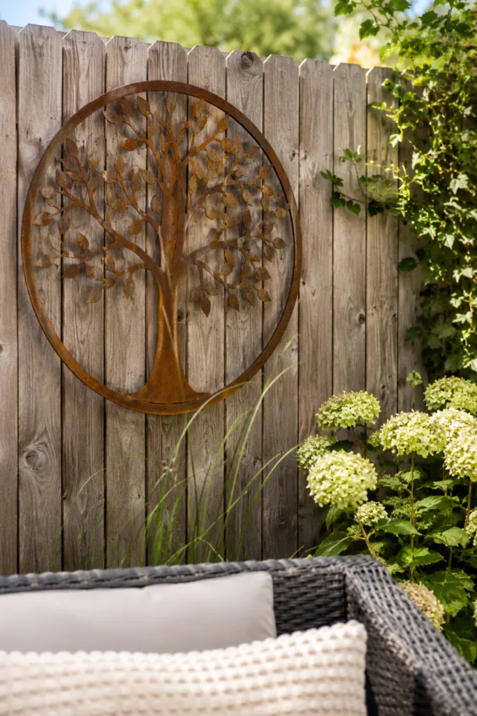 Sommerdeko für den Gartenzaun – 5 einfache Ideen für den Juli Gartenzaun Deko im Juli mit rostfarbenem Metallobjekt an Holzzaun im sommerlichen Garten.