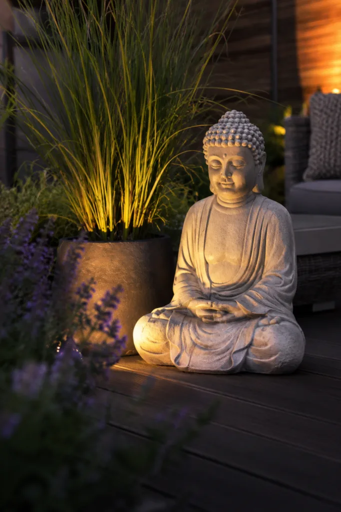 Terrassendeko mit Figuren: Beleuchtete Steinfigur auf Terrasse zwischen Gräsern und Pflanzen am Abend.