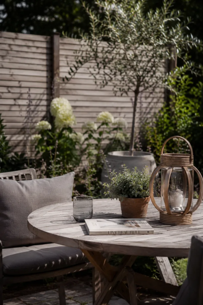 Hintergartendeko mit kleinem Sitzplatz, Holztisch, Laterne und Pflanzen im ruhigen Garten.