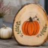 Herbstdeko basteln für den Garten mit bemalter Holzscheibe, Heide, Zapfen und kleinen Kürbissen.