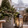 Winterdeko für den Balkon mit Tannengrün im Kübel, großer Laterne, Lichterkette und verschneitem Hintergrund.