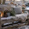 Winterdeko für deine Gartenbank mit Laterne, Kissen und Zweigen im verschneiten Garten.