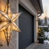 Großer beleuchteter Weihnachtsstern aus Metall und Glas an einer Steinwand neben einer verschneiten Garageneinfahrt.