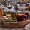 Holztablett mit Glühwein, Weihnachtsplätzchen, Tannenzweigen und Laterne auf einem winterlichen Gartentisch.
