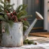Alte Zinkgießkanne mit Tannenzweigen, roten Beeren und Zapfen als Weihnachtsdeko vor einem Hauseingang.