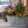 Große Zinkwanne mit Tannengrün, kleiner Konifere, Zapfen und warmweißer Lichterkette auf einer winterlichen Terrasse.