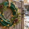 Weihnachtskranz aus altem grünem Gartenschlauch mit Tannenzweigen, Zapfen und Lichterkette an einem Holztor.