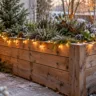 Weihnachtliches Hochbeet aus Holz mit warmweißer Lichterkette, Tannengrün und Laterne auf verschneiter Terrasse.