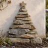 Weihnachtsbaum aus übereinandergestapelten flachen Natursteinen vor einer hellen Gartenmauer.