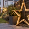 Großer beleuchteter Metallstern vor einem dunklen Holzzaun mit immergrünem Pflanzkübel im Reihenhausgarten.